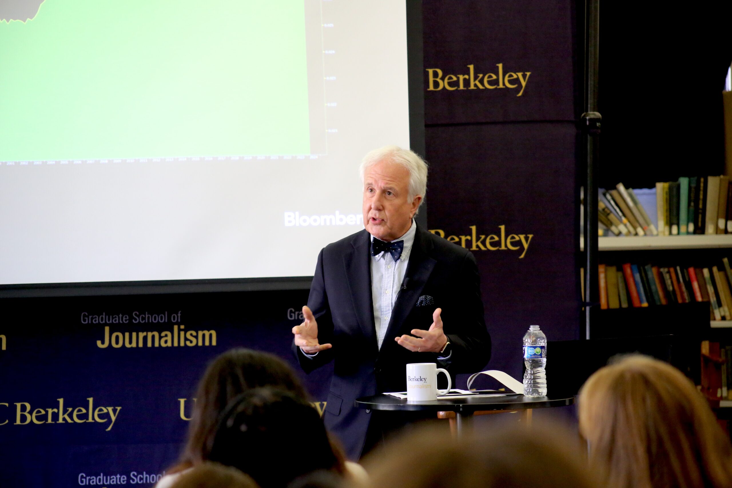 Man in a blue suit with bow tie and white hair speaks with students.