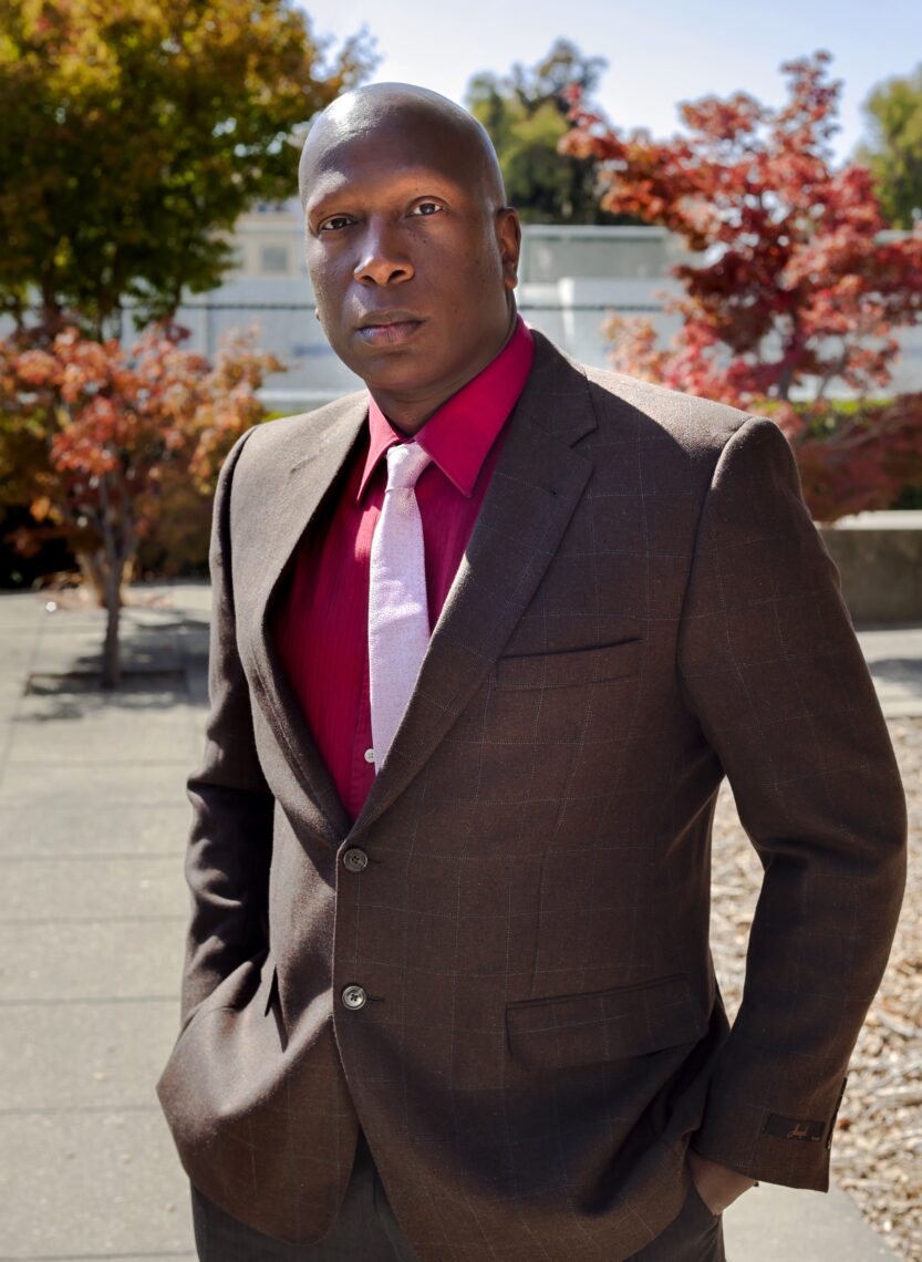 A tall bald African American man in a beautiful brown suit, red shirt and pink tie with trees in the background.