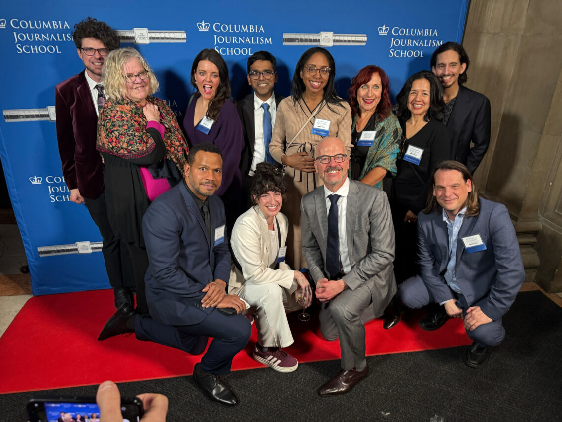 Group image of journalists from Reveal on the red carpet in front of a blue duPont Awards banner smiling.