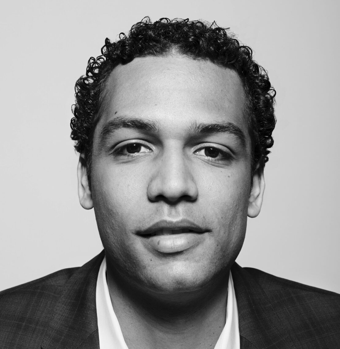 Closeup black and white portrait of an African American man with short curly black hair wearing a suit in front of a gray background.