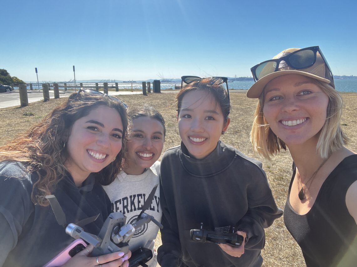 Three female students stand close to their instructor with blonde hair wearing a baseball hap in front of a field.