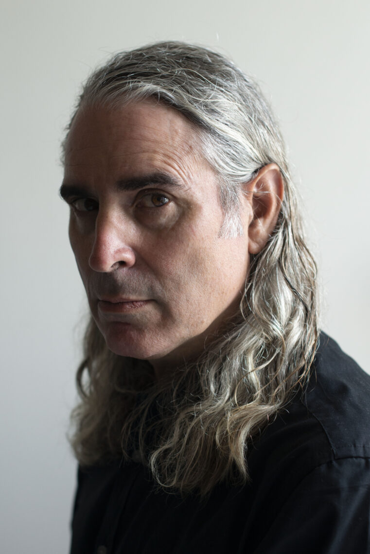 Man with long gray wavy hair wearing a black turtleneck and looking intently at the camera from profile.