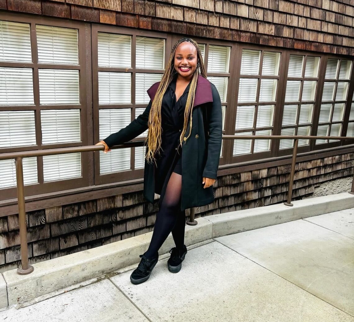 A young woman with long hair and dressed in black jacket and skirt and boots smiles while holding a railing in front of a brown shingled building at North Gate Hall.