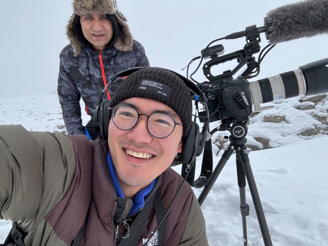 Two mean smiling at the camera while filming in Pakistan with snow and film gear in the background.