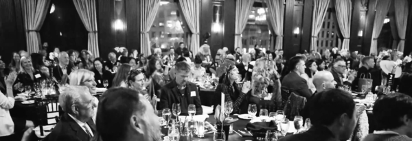 Reporters sit at round tables with tablecloths/dinner settings at an awards ceremony with curtained windows in the background.