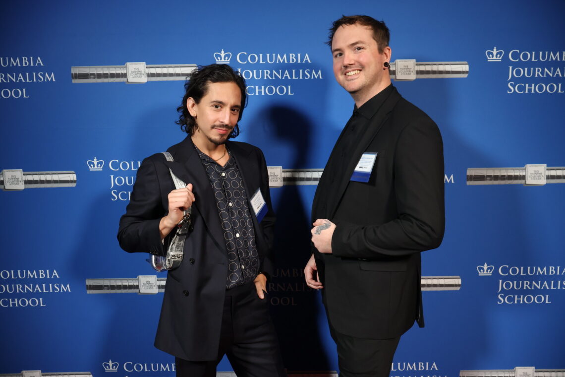 Two mean dressed in black suits on the red carpet smiling with a blue background with white and silver duPont Awards logos.