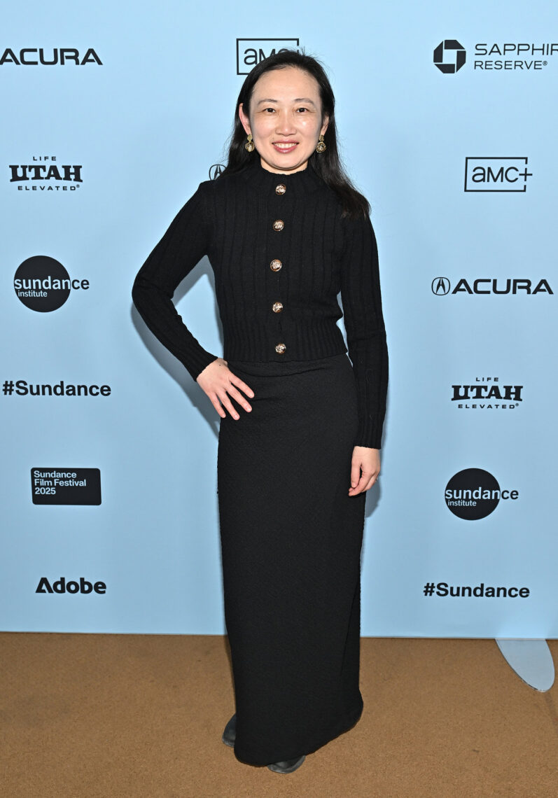 An Asian woman wearing a long dark suit dress with her hand on her hip on the carpet at the Sundance Film Festival.