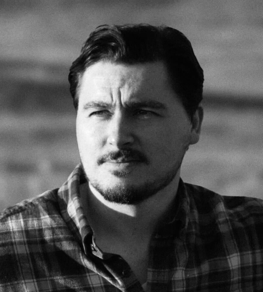 A handsome young reporter wearing a plaid button down shirt with dark hair and a mustache with his head turned to the side with sun on his face.