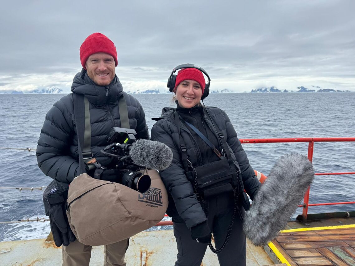 A man and woman on a boat with mountains and open water in the background holding camera gear smiling.