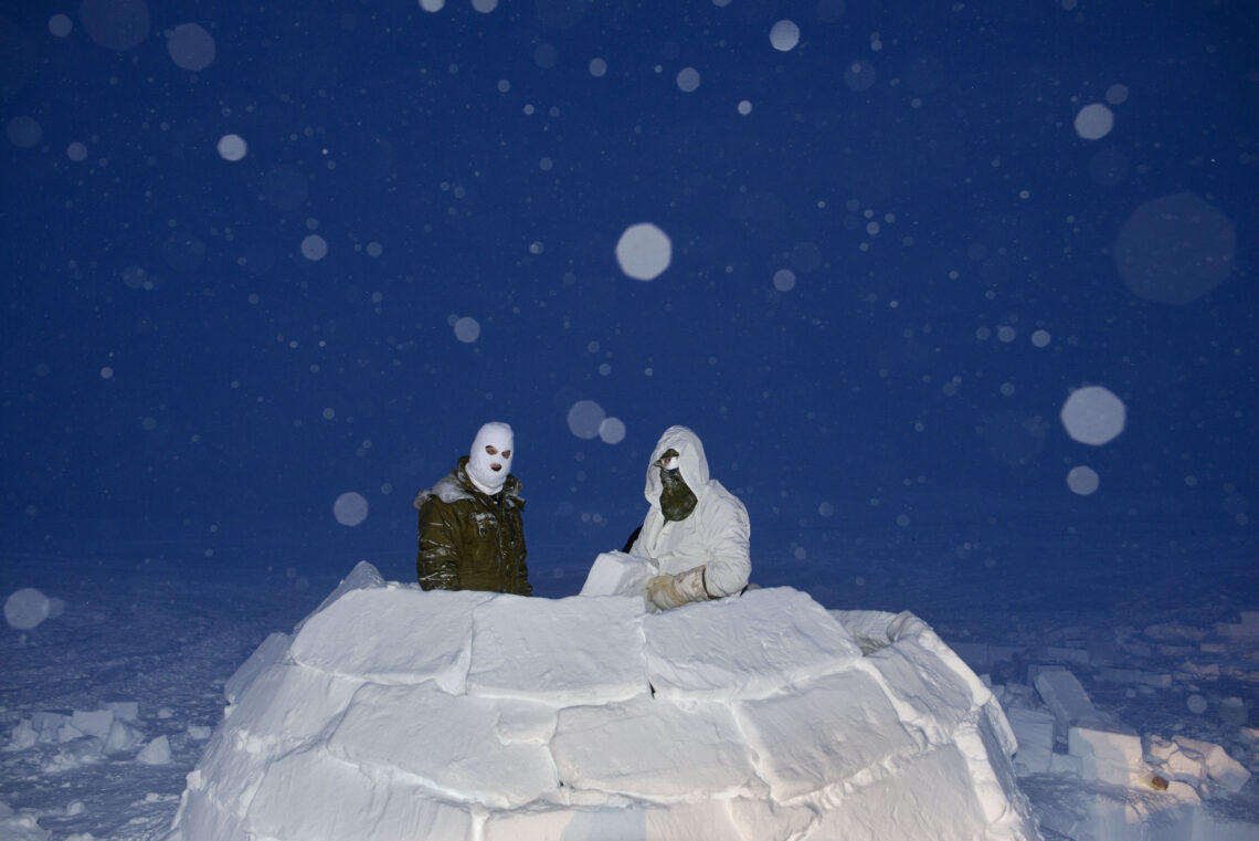 Two people wearing white winter gear on top of an igloo with a dark sky in the background.