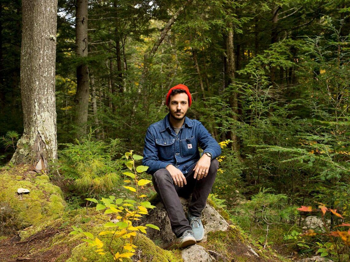 A man with brown hair wears a red knit beanie and a denim shirt sitting on a log in the woods.