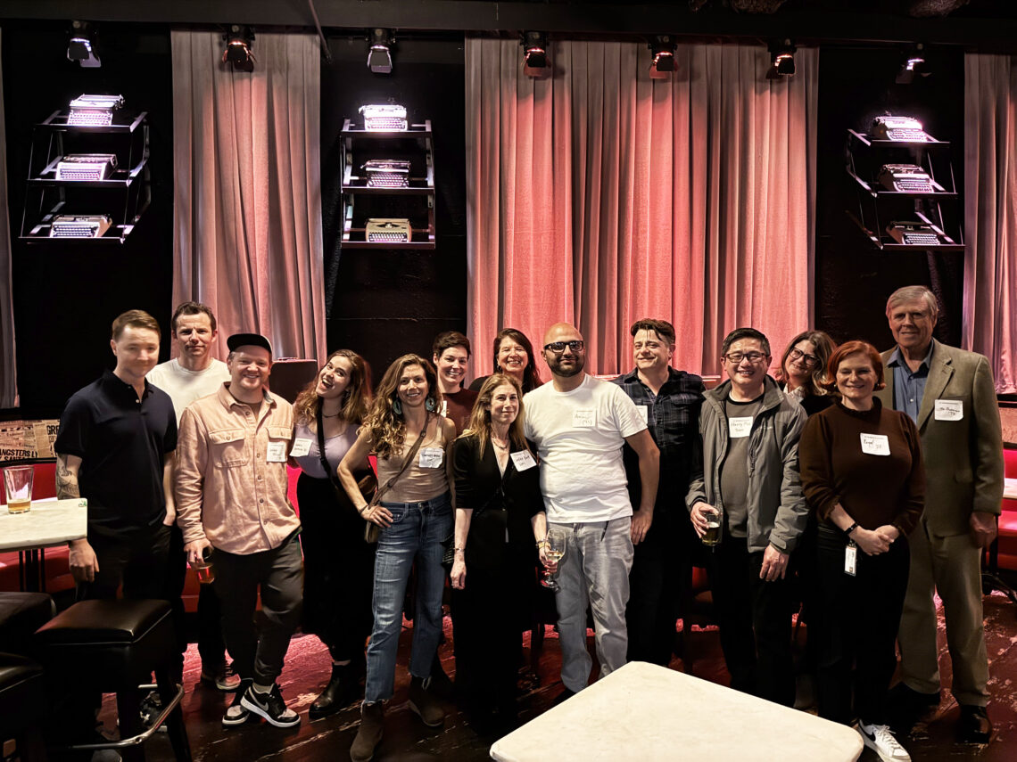 A large line of journalists stand next to eachother in a restaurant with red lights in the background.