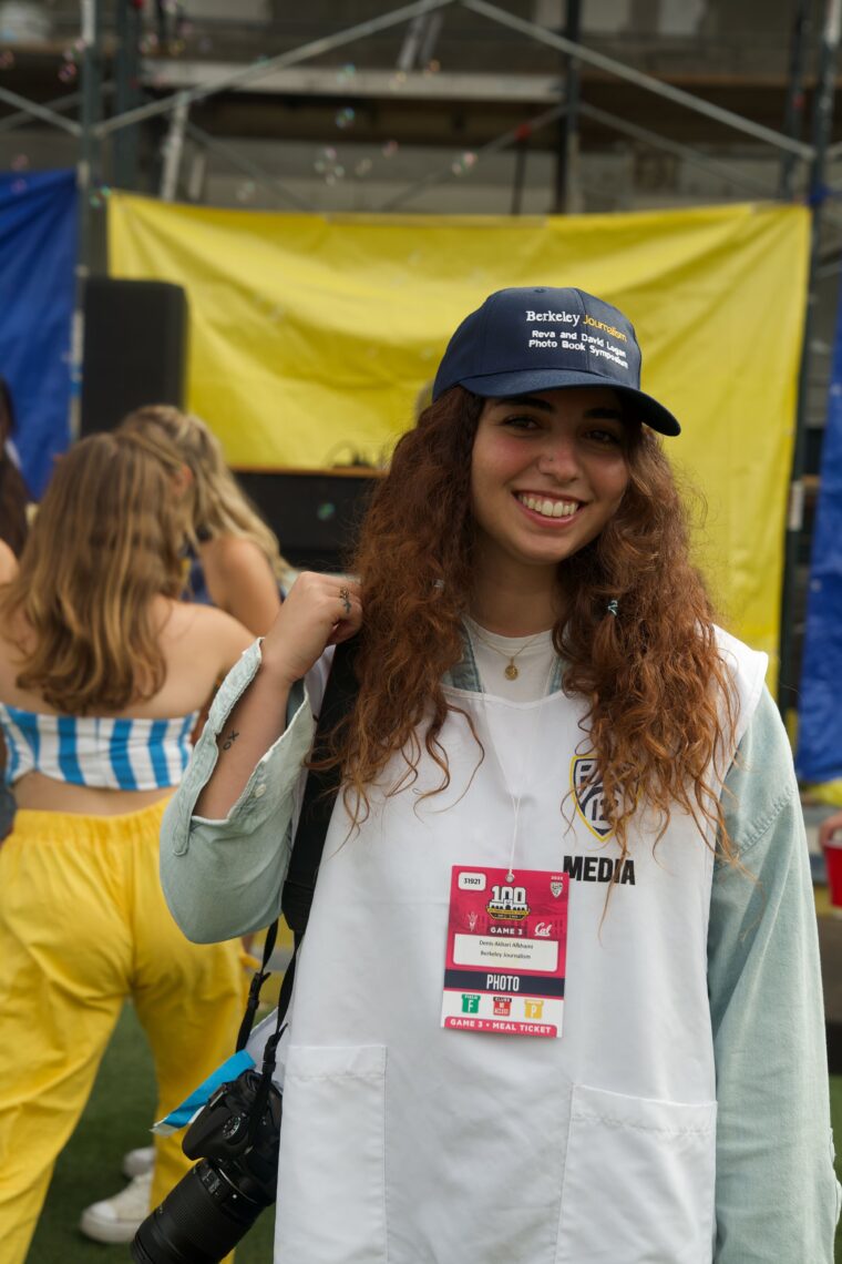 A young woman with long brown wavy hair wears a press pass and hat smiling.