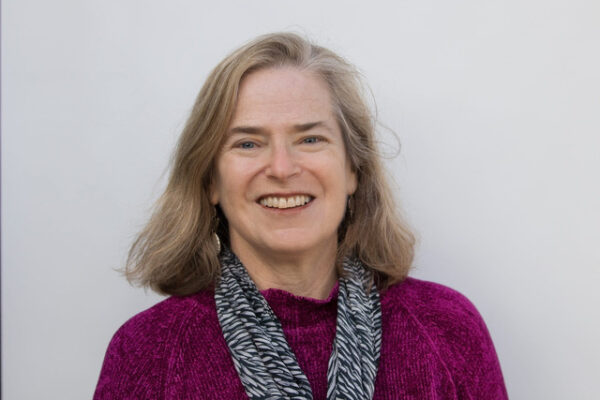 A woman with shoulder length dark blonde hair wearing a burgandy sweater and black and white printed scarf smiling in front of a light gray background.