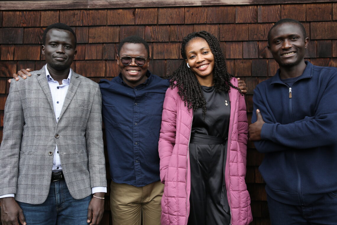 Four graduate students, one woman and three men, stand next to oneanother smiling in front of a brown shingled building.