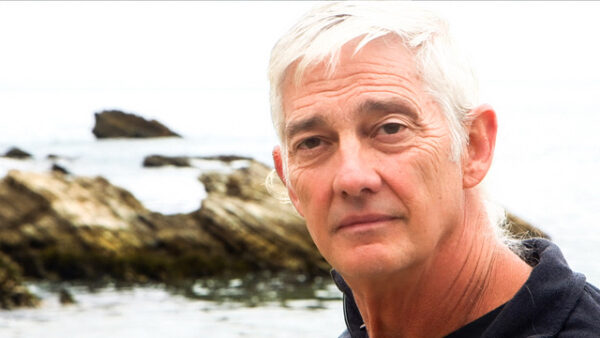 head of a man with white hair standing in front of a shoreline.