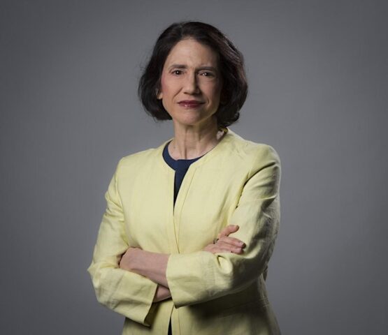 woman with brown hair and red lipstick with a yellow suit jacket, folding her arms and looking seriously at the camera