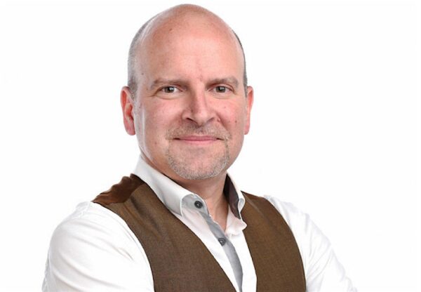 Bald man in a white shirt and brown vest looking serious but happy