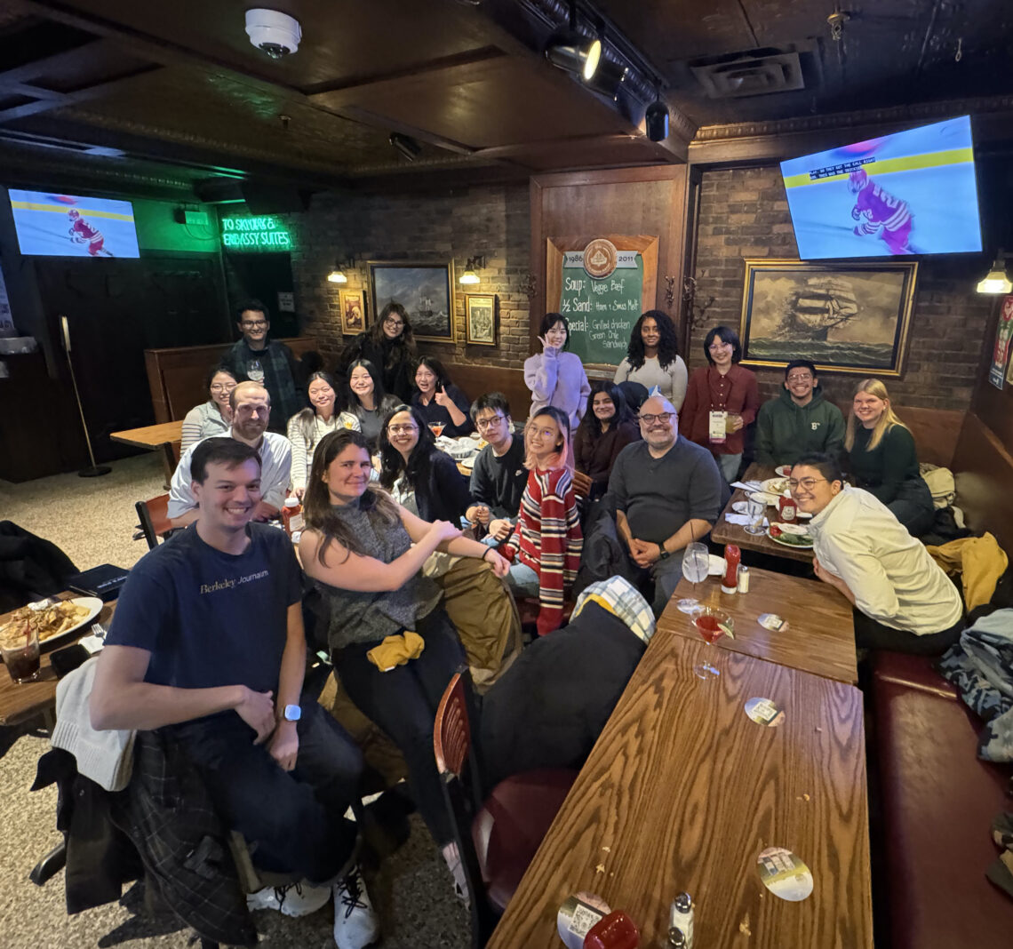 Large group of young data journalists sit in a restaurant surrounded by TV screens smiling.