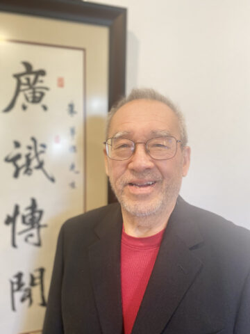 Man in a black sports coat and red t-shirt against a screen with Chinese characters. 