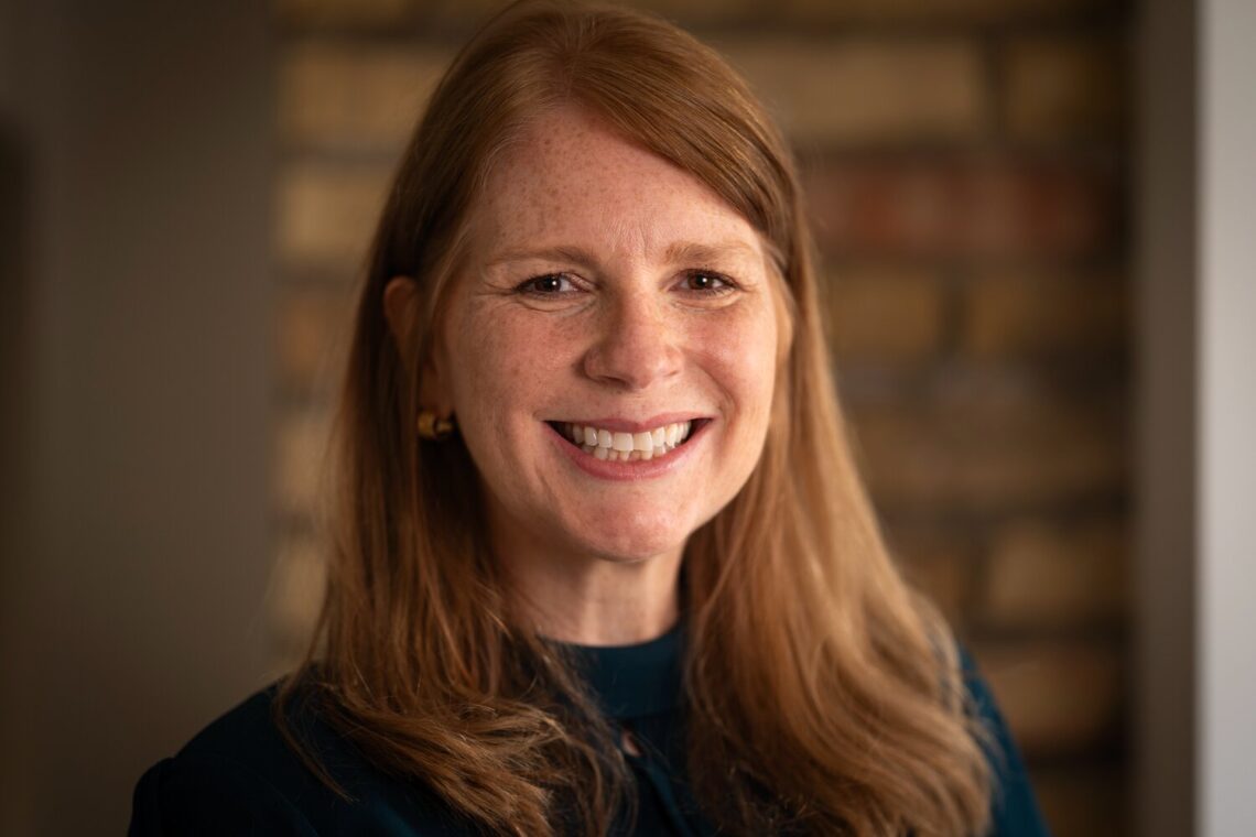 A woman with reddish shoulder length hair photographed in front of a brick background smiling brightly.