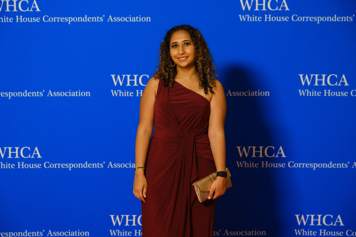 A young woman with brown curly shoulder length hair wears a beautiful plum colored one shoulder dress in front of a blue WHCA background.