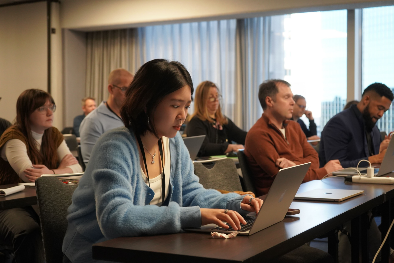 Young woman types at a computer in a room with others who are looking ahead at a speaker. Some have computers. They are various ages and ethnicities.
