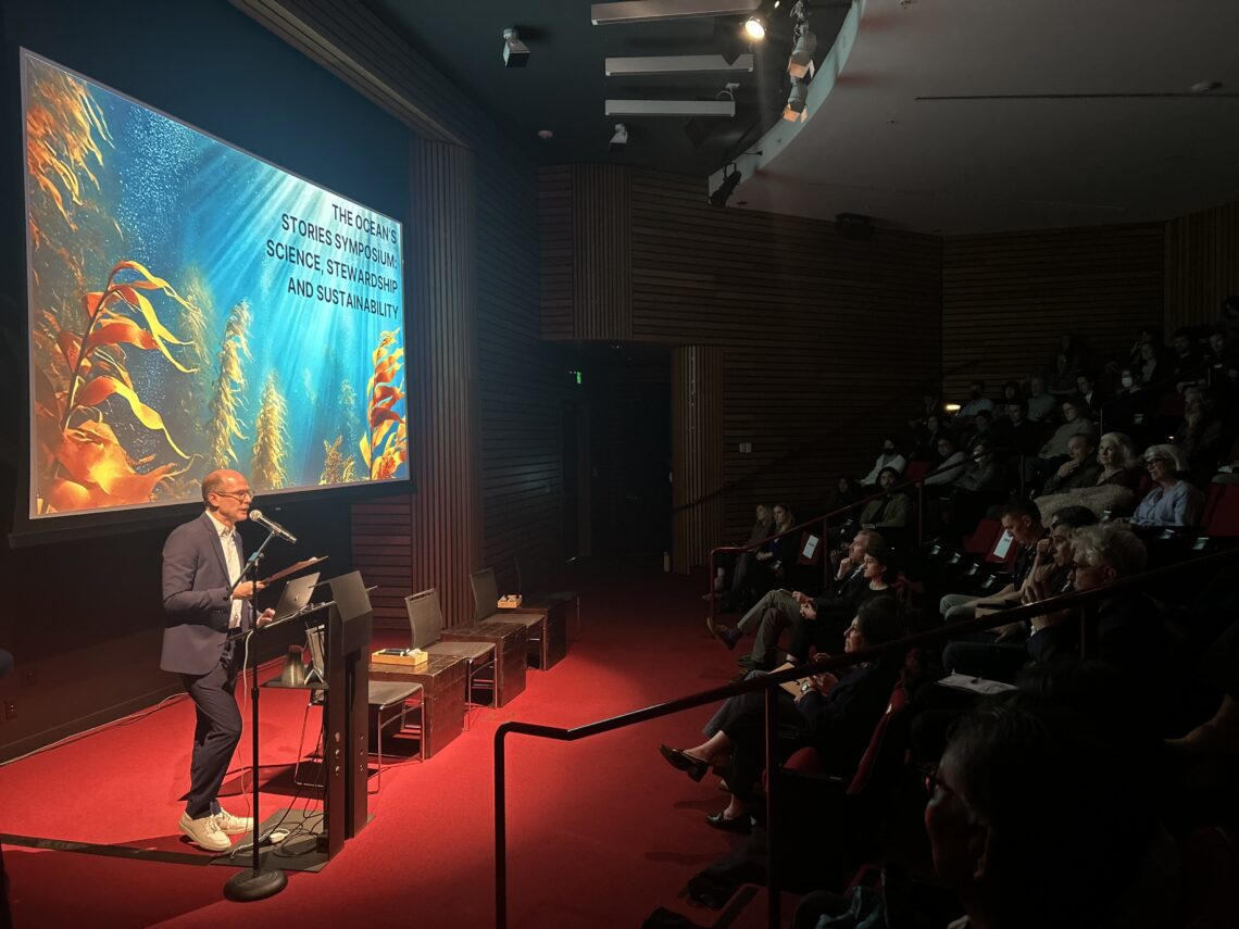 Man on a stage with red carpet and image of an ocean behind him.