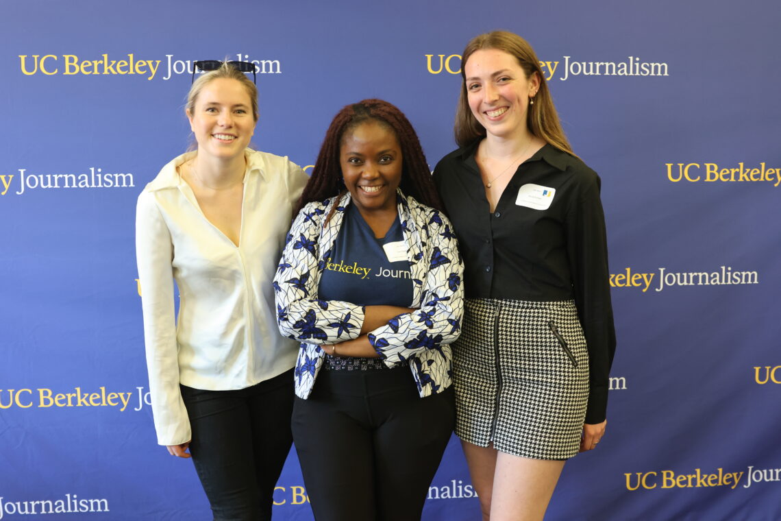 Three women stand in front of a blue Berkeley Journalism banner smiling.