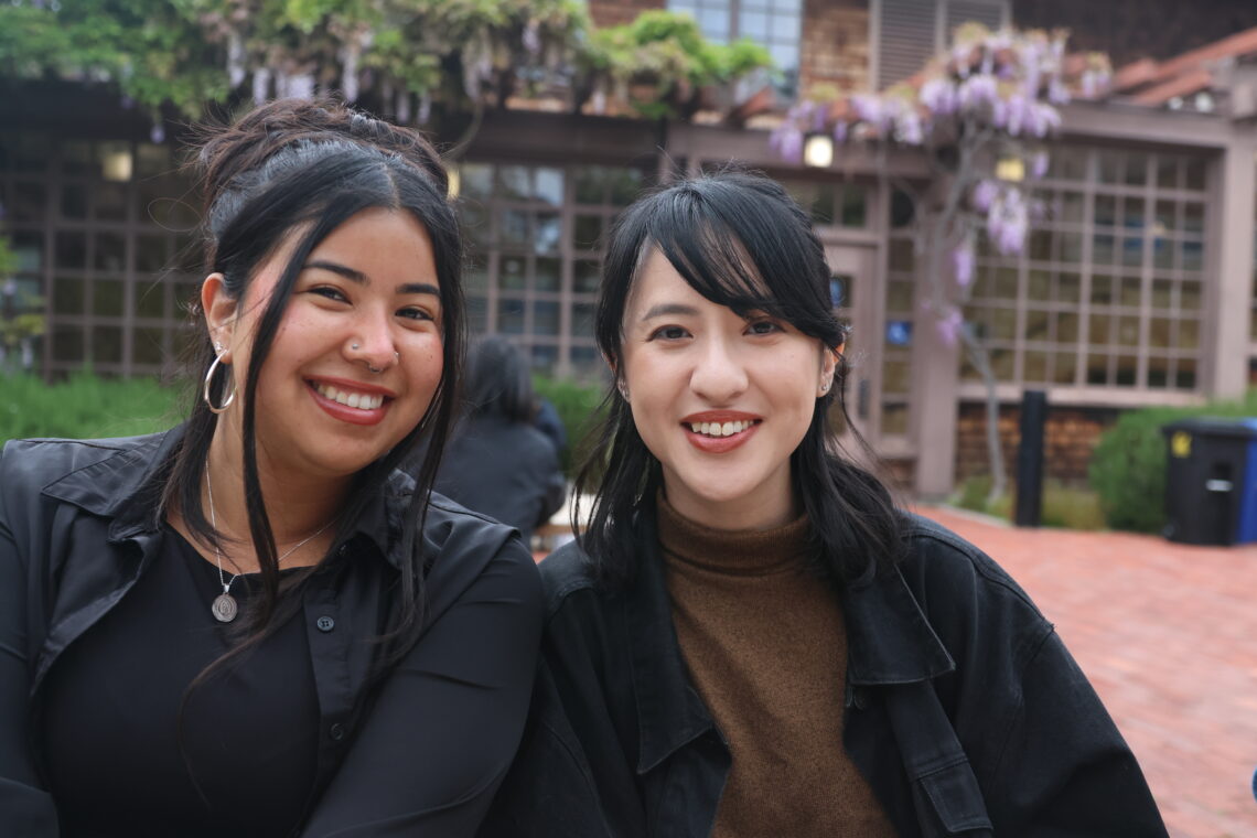 Two women graduate students sitting close to oneanother in front of a brown shingled building with purple wisteria flowers smiling brightly.