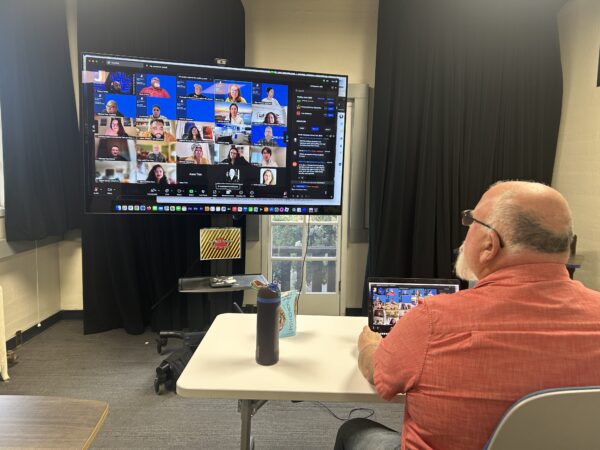 A man wears a short sleeve coral colored shirt seated at a white table monitoring a large Zoom meeting on a color TV screen.