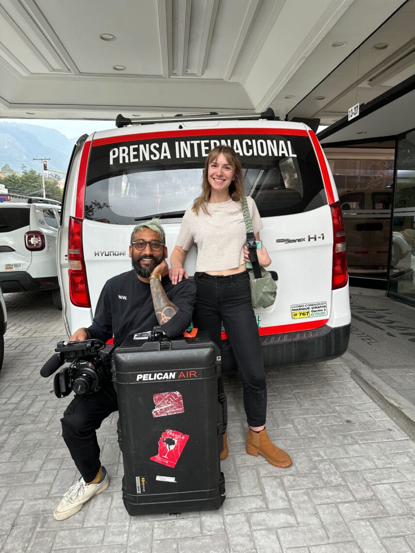 A man and woman sit on film suitcases and the bumper of a white van parked at an airport smiling.