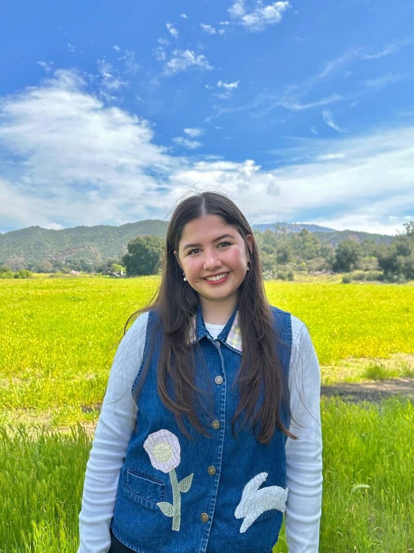 A young woman with long dark hair stands in a bright yellow field with hills in the background smiling.