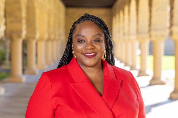Woman in a red/orange suite in a long hallway with a slight smile.