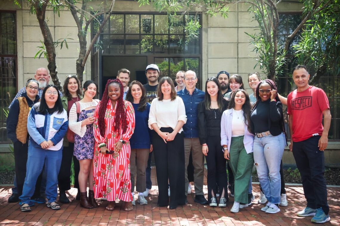 A large group of graduate students and their instructors standing in front of a building with glass and metal doors smiling.