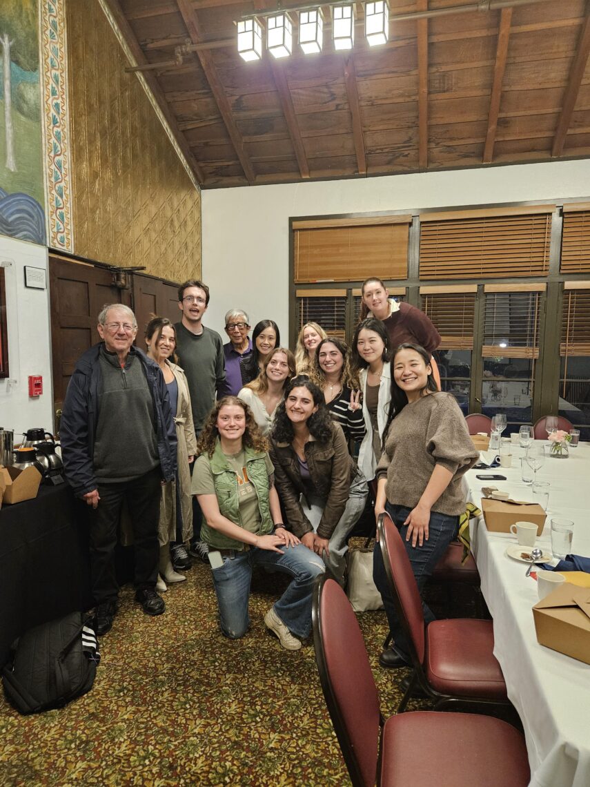 A group of a dozen students stand next to their professor near a restaurant table.