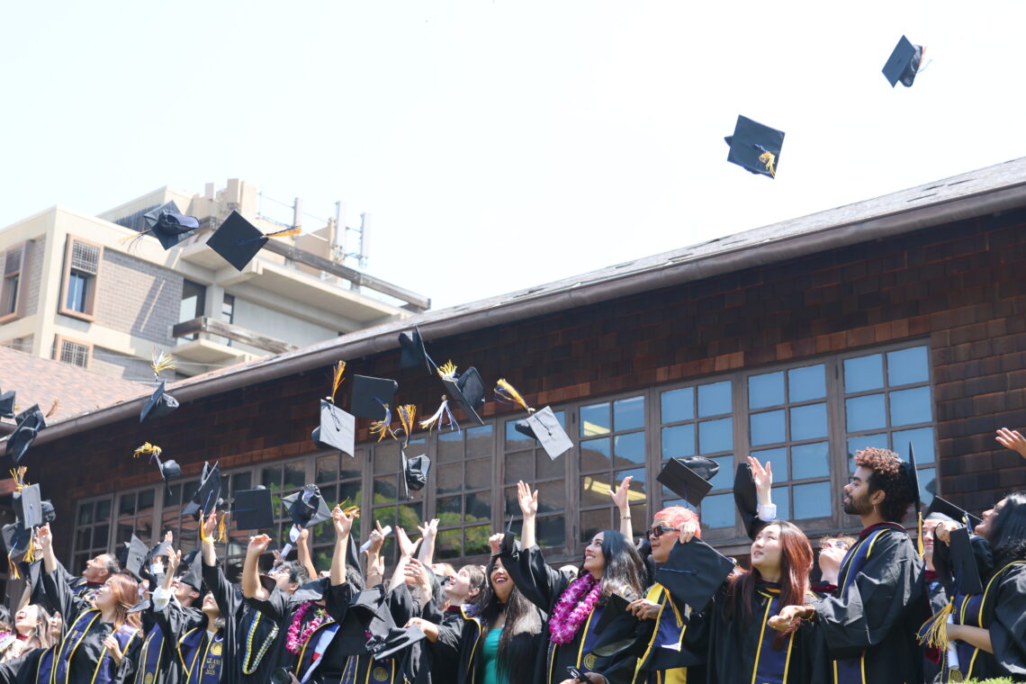 Photo of a large group of students in gowns throwing their caps in the air in front of a brown shingled building.