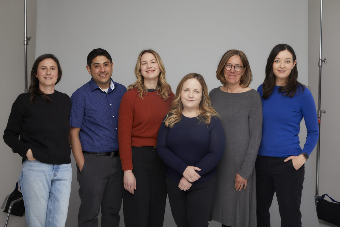 A group of 6 journalists stand in front of a white wall.