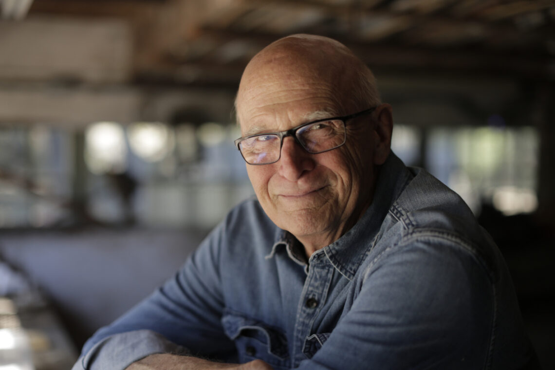 An older filmmaker photographed in semi-profile wearing glasses and his arms crossed on a table in dramatic light.