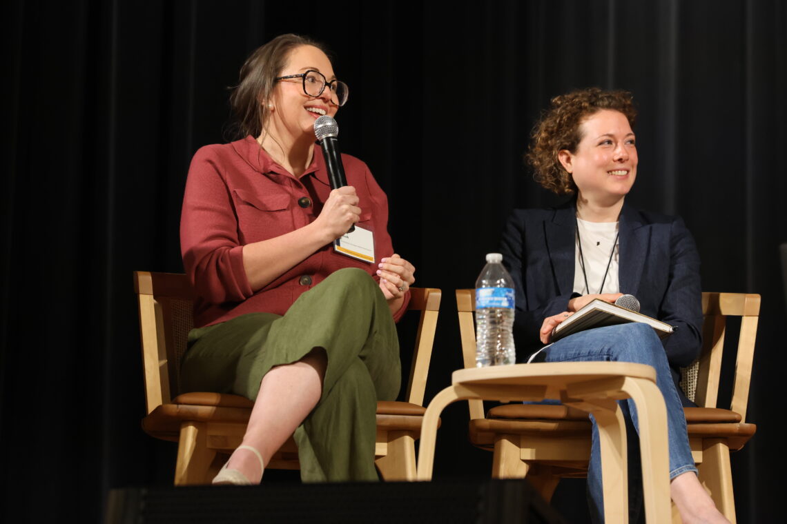 Two women speaking into microphones on a stage dressed in business casual clothes.
