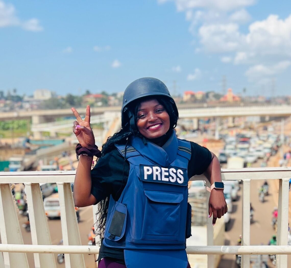 A beautiful African woman wears a blue press security vest and blue helmet leaning against a white raining giving the peace sign smiling.