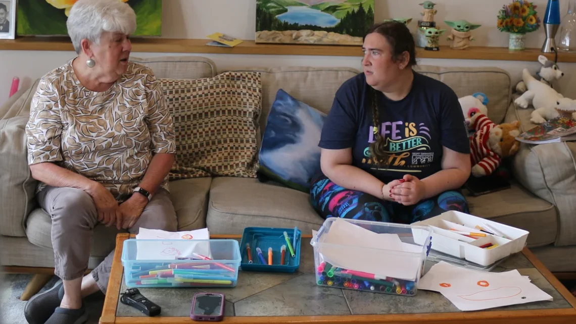 Rachel and Linda Tung sit on a beige couch, looking at one another. In front of them is a coffee table with paper and markers.