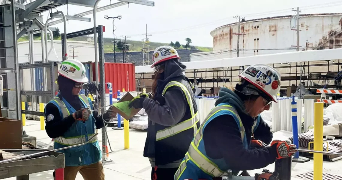 Three refinery workers dressed in safety gear and hardhats working.