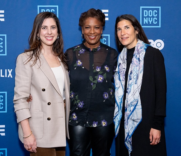 Three women stand next to eachother in front of a DocNY blue banner with logos smiling.