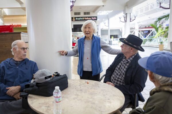 Three older men sit at a round table, one older woman is speaking to them and gesticulating. 