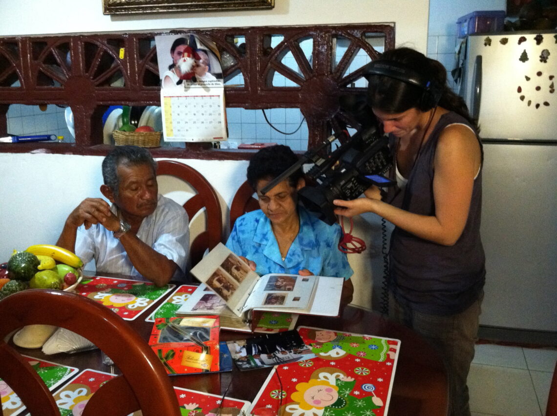 A woman holding a film camera films two women at a kitchen table.