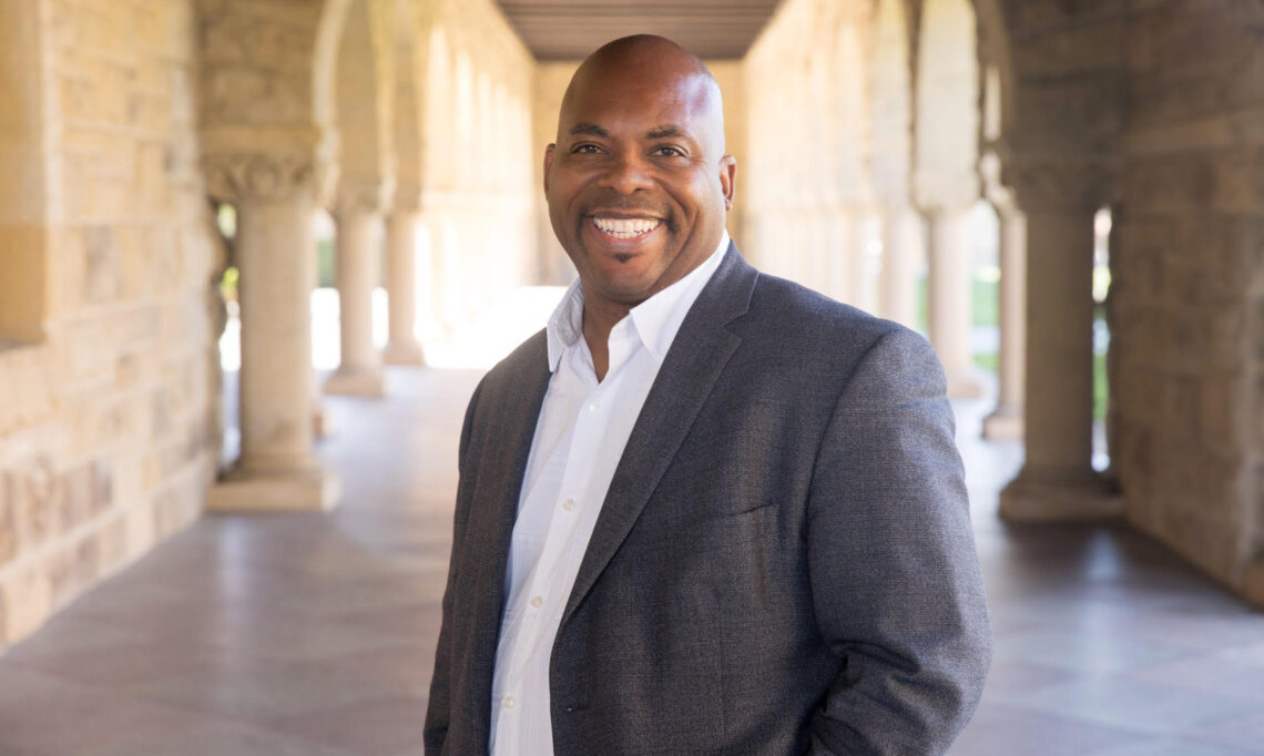 An African American man wearing a black suit jacket over a white button down dress shirt stands in front of a descending row of white exterior columns smiling.