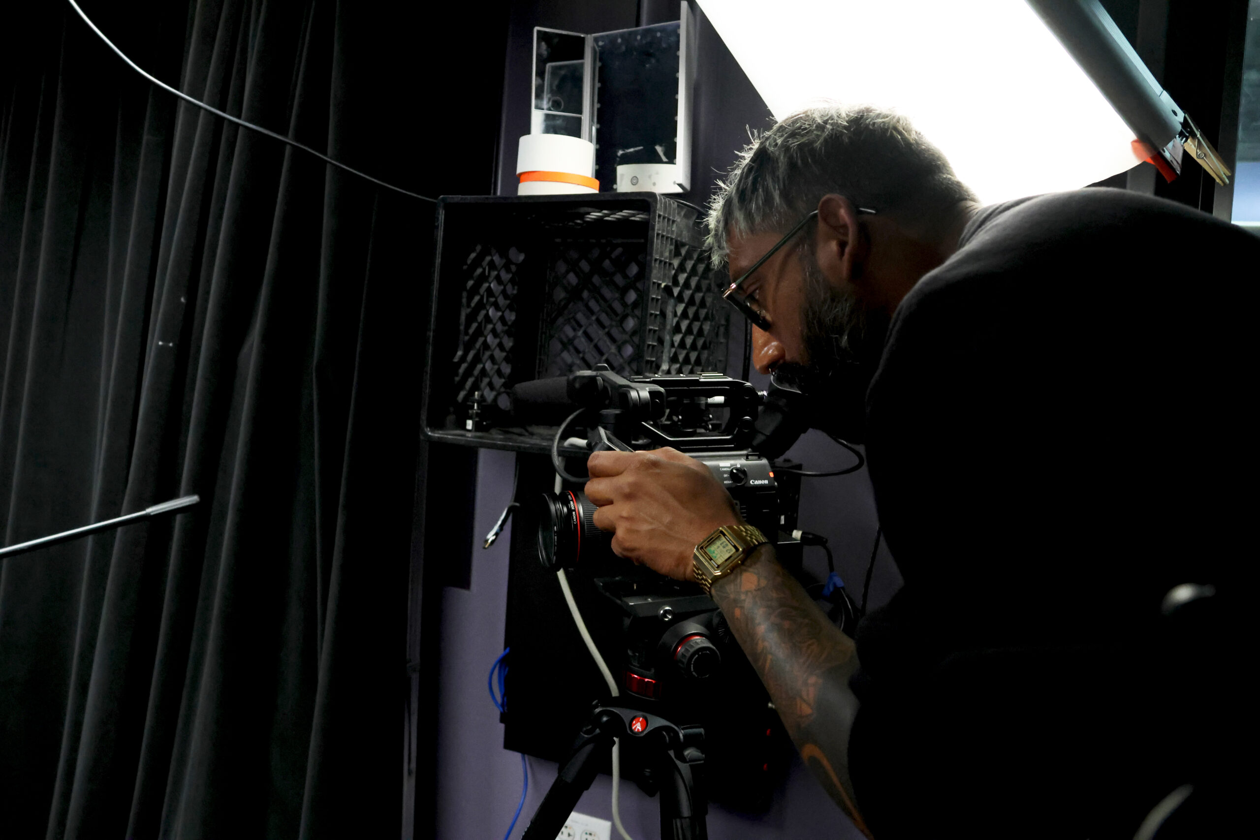 A man filming in a dark studio with a white square lamp above him. His arm has tattoos.