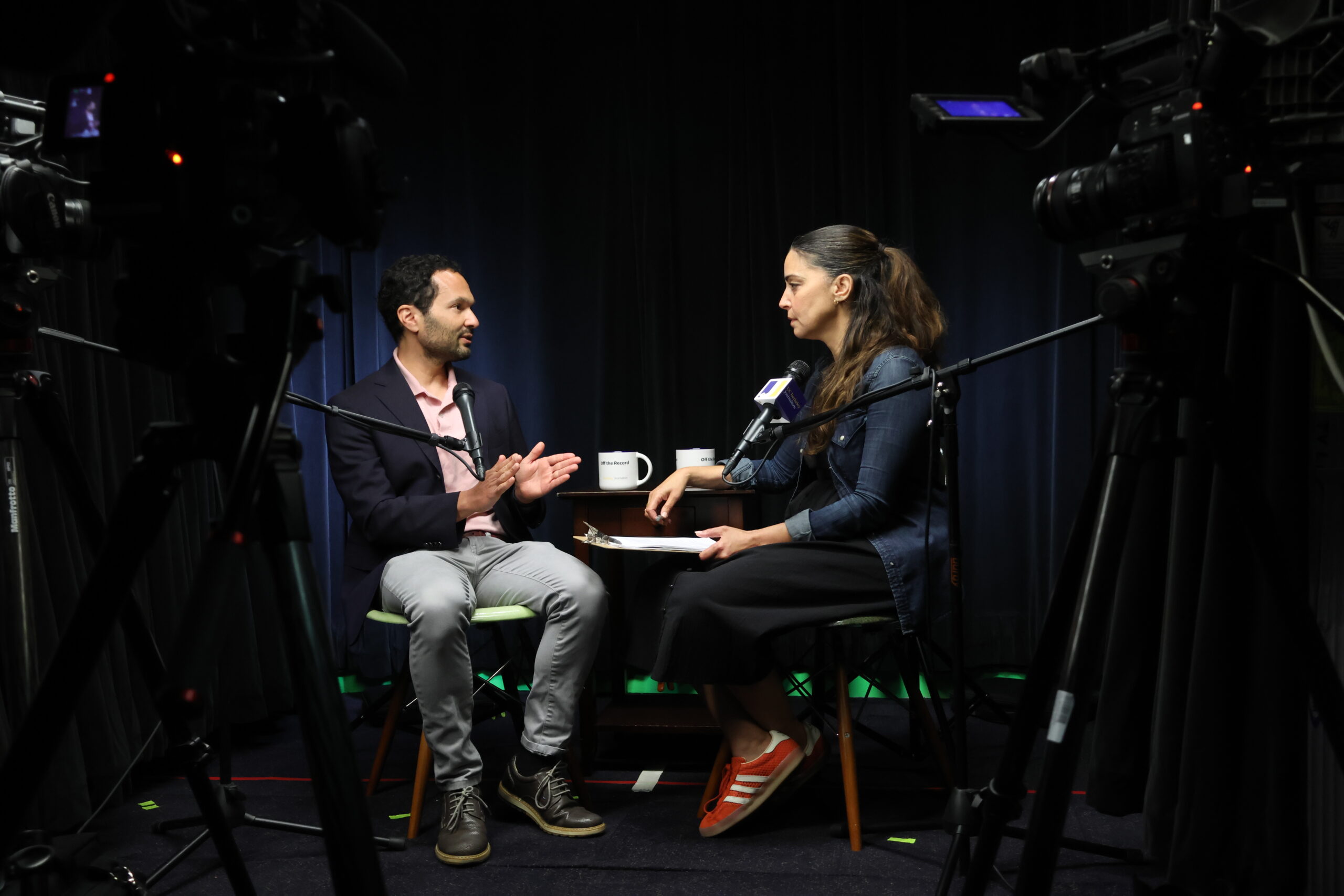 Two people doing an interview facing each other in chairs in a dark studio with camera equipment.