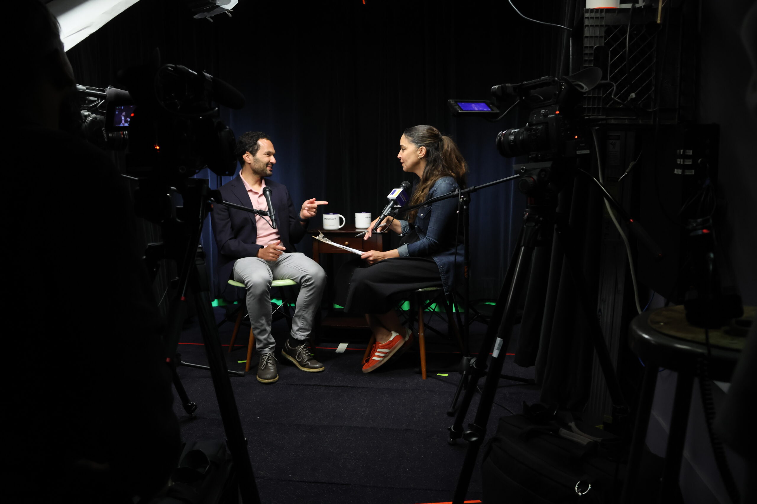 Two people doing an interview facing each other in chairs in a dark studio with camera equipment.